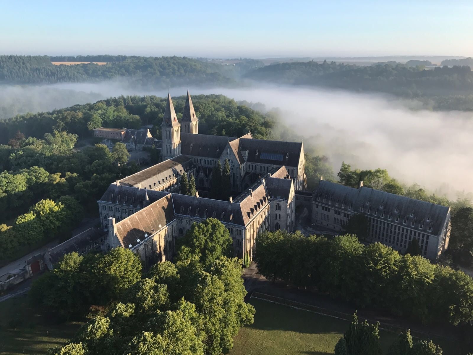 Vue aérienne de l'abbaye de Maredsous au petit matin, ses deux flèches dressées au-dessus des arbres, la vallée de la Molignée noyée dans la brume.